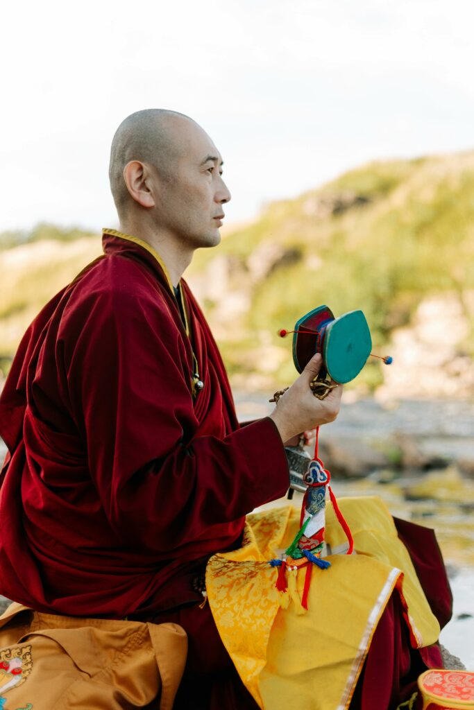 A Buddhist monk in traditional robes sits in meditation with a Tibetan prayer wheel outdoors.